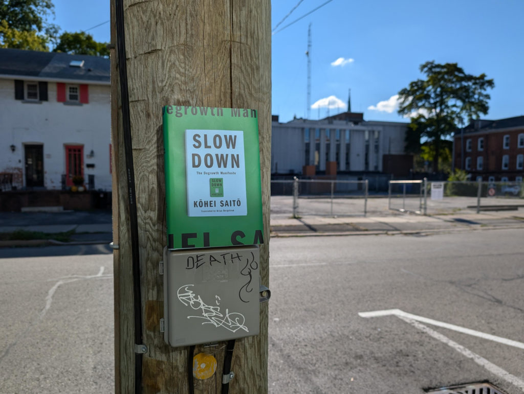 Photo of a hardcover copy of Slow Down sitting atop some kind of grey plastic electrical box with thick black wires coming in and out of it on a wooden telephone pole. Some one has written "DEATH" on the box in permanent marker and it's been tagged a few times. Behind the pole is an urban street and a variety of residential-looking buildings. 