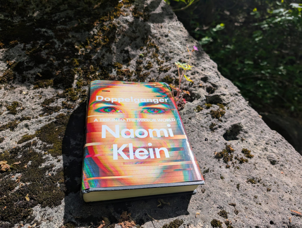 My hardcover copy of Doppelganger basking in the sun on a moss-spattered rock with a small plant flowering out of the top right corner. The sun hitting the glitchy cover like the way it is here makes the red look especially unsettling and sinister.
