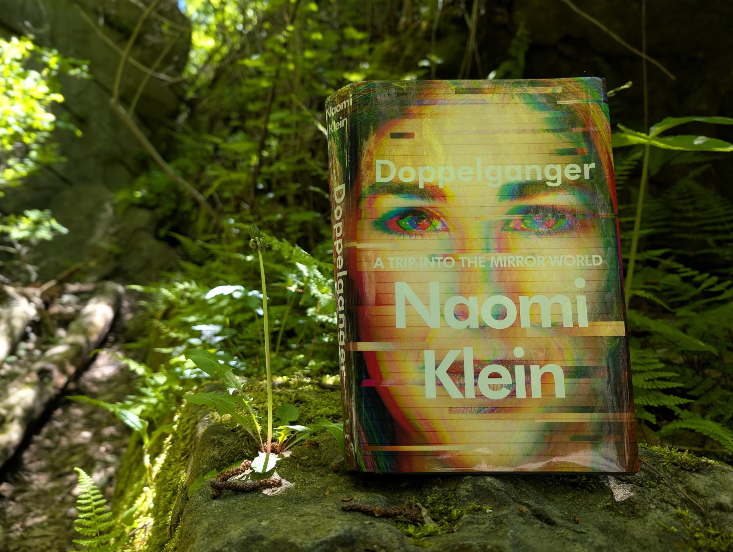 Picture of my beat up copy of Doppelganger. It is hardcover and features a freaky, glitchy picture of Klein's face in red, green, and the original colours with the title, subtitle, and author's name in white. The book is standing on a moss-covered rock beside a dandelion deflowered and in a rocky green wood.